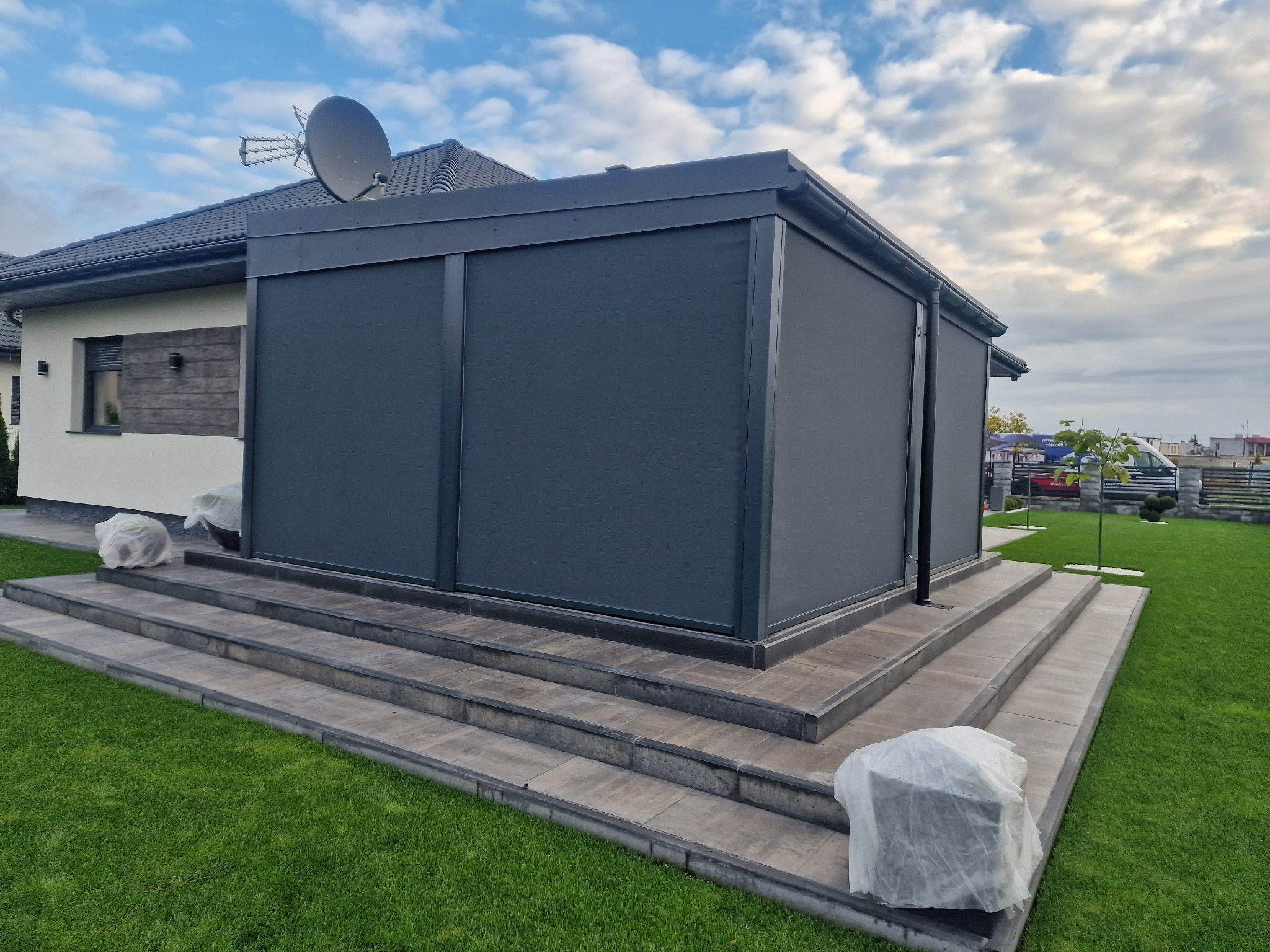 Zadaszony taras z szarymi roletami zewnętrznymi, widok z poziomu trawnika, częściowo zasłonięte meble ogrodowe na schodach.