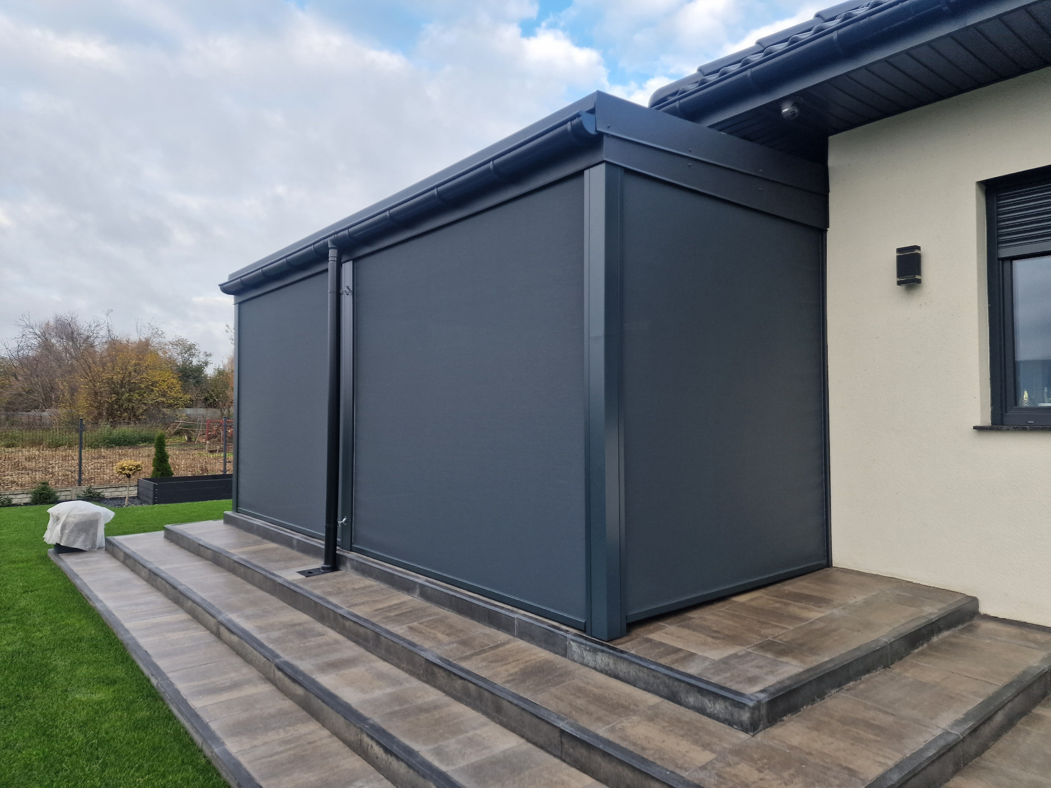 Zewnętrzna zabudowa tarasu z grafitowymi roletami pionowymi, widok z boku na schody wejściowe i fragment ogrodu.