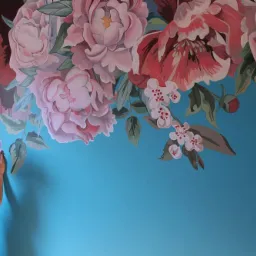 Kobieta w kwiecistej koszuli maluje na niebieskiej ścianie mural z dużymi, różowymi kwiatami piwonii i gałązkami kwitnącej wiśni.