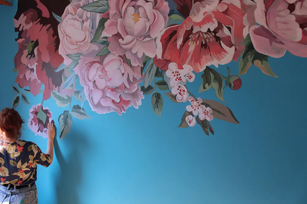 Kobieta w kwiecistej koszuli maluje na niebieskiej ścianie mural z dużymi, różowymi kwiatami piwonii i gałązkami kwitnącej wiśni.