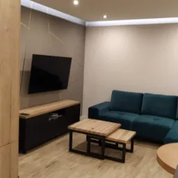 Jasne panele podłogowe w salonie z turkusową sofą narożną, niskim stolikiem kawowym z drewna i metalu, oraz szafką RTV z wiszącym telewizorem na ścianie z geometrycznym wzorem.