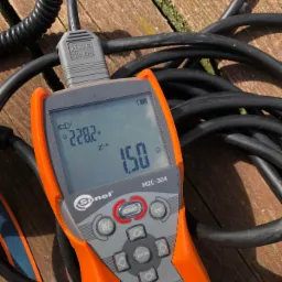 Miernik Sonel MZC-304 wyświetlający wynik pomiaru impedancji pętli zwarcia 1.50 Ω, leżący na drewnianym podłożu obok czarnego przewodu pomiarowego.