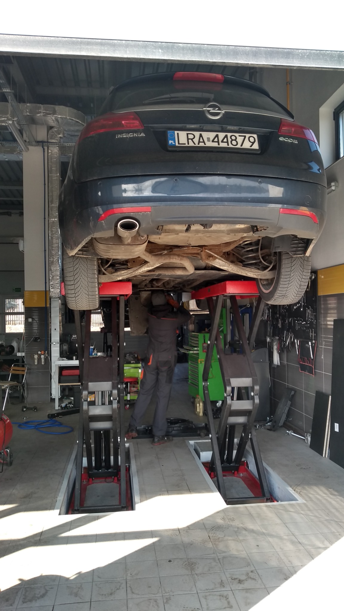 Opel Insignia kombi na podnośniku dwukolumnowym w warsztacie, widok od spodu pojazdu podczas pracy mechanika w szarym kombinezonie. Widoczne narzędzia i wyposażenie warsztatu.