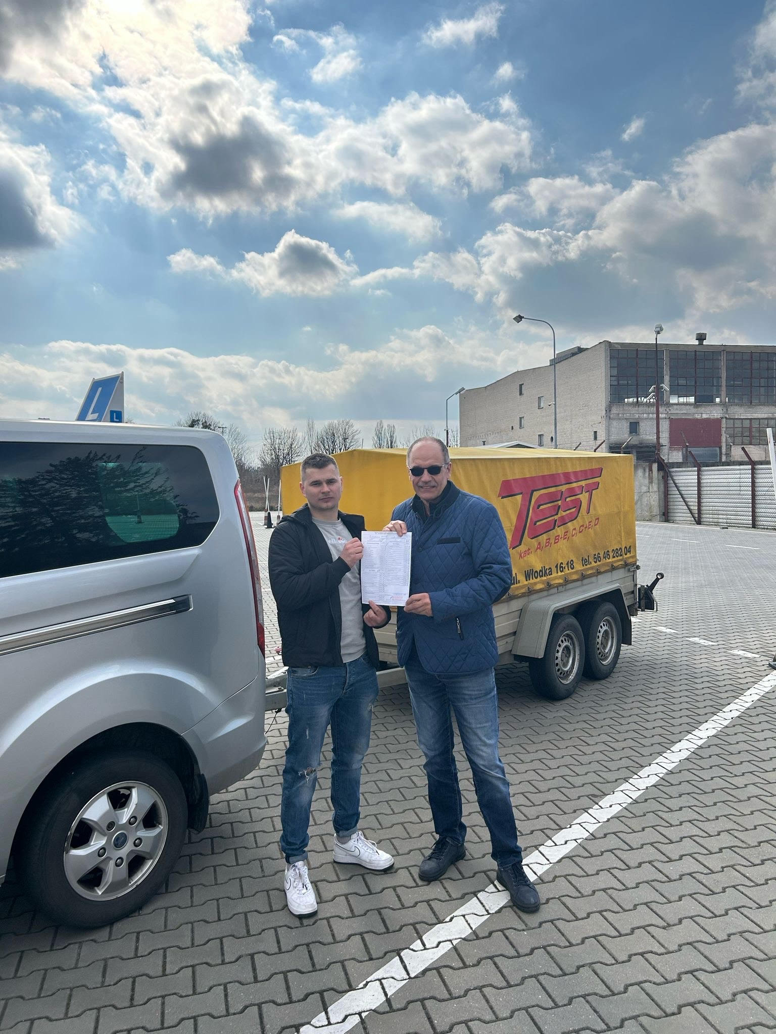 Dwaj mężczyźni stojący obok srebrnego vana i żółtej przyczepy z napisem 'TEST' oraz informacjami o kategoriach prawa jazdy, trzymający dokumenty na parkingu z kostki brukowej.