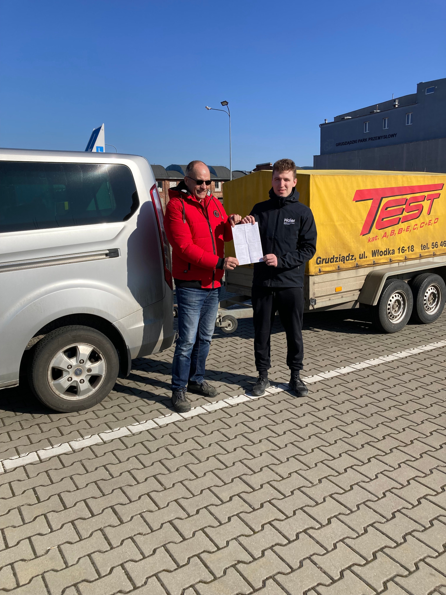Mężczyzna w czerwonej kurtce przekazuje dokument młodemu mężczyźnie w czarnej kurtce na tle srebrnego vana i żółtej przyczepy z napisem 'TEST' i informacjami o kategoriach prawa jazdy,...