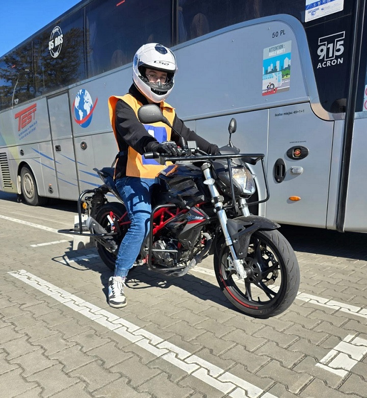 Instruktor w kasku i kamizelce odblaskowej siedzi na czarnym motocyklu marki Benelli, zaparkowanym przed autobusem z naklejkami informacyjnymi.