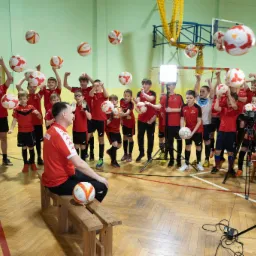 Grupa młodych piłkarzy w czerwonych strojach pozuje z piłkami w hali sportowej podczas sesji zdjęciowej, oświetlonej lampą i filmowanej kamerą na statywie.
