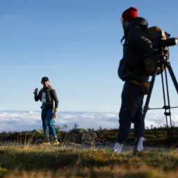 Fotograf z kamerą na statywie filmuje sportowca na tle górskiej panoramy z chmurami, w słoneczny dzień.