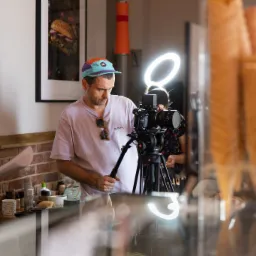 Ekipa filmowa kręci ujęcie w kawiarni, widoczny kamerzysta z profesjonalną kamerą na statywie i lampą pierścieniową, asystent z dokumentami oraz ekspozycja lodów w wafelkach w szklanej witrynie...