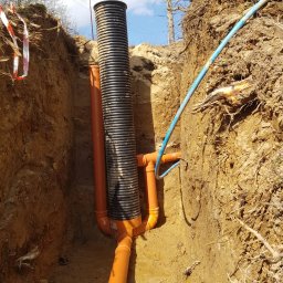 Usługi budowlane - Głęboki wykop z ułożonym systemem rur drenarskich w Mirsku. Widoczne pomarańczowe rury połączone z czarną rurą karbowaną. Nad wykopem wystają korzenie drzew.