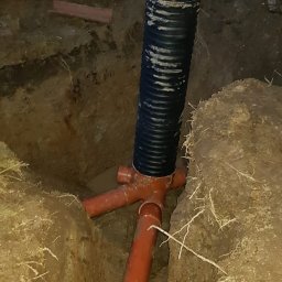 Usługi budowlane - Instalacja drenażowa z rurą karbowaną w wykopie. Widoczne złącze trójnikowe i fragmenty rur. Ziemia wokół wykopu, słabe oświetlenie.