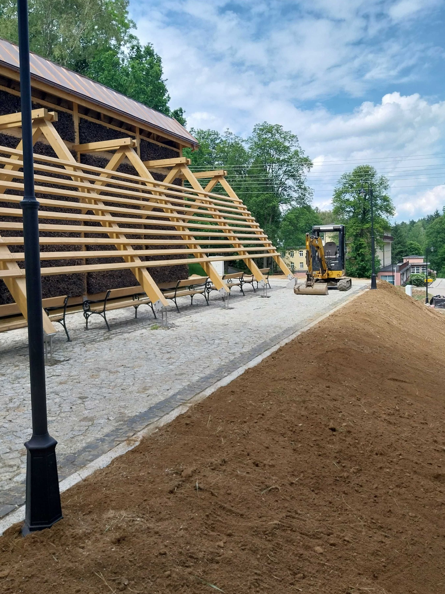 Budowa drewnianej konstrukcji solankowej z mini koparką w tle, świeżo usypana ziemia, park miejski w słoneczny dzień.