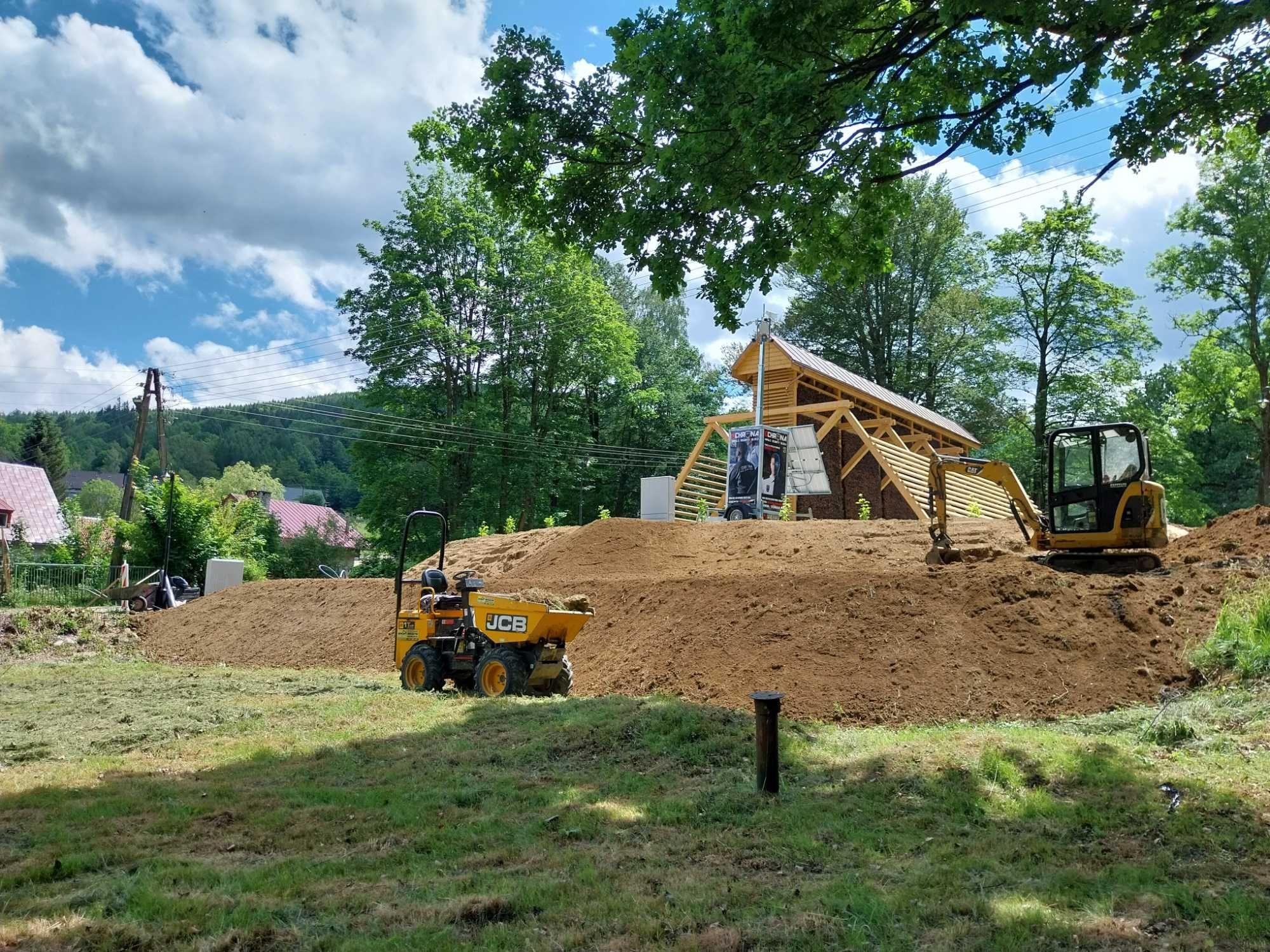 Żółta mini wywrotka JCB na trawiastym terenie, w tle koparka i budynek w trakcie budowy z drewnianą konstrukcją dachu, ziemia usypana w kopiec. Słoneczny dzień.