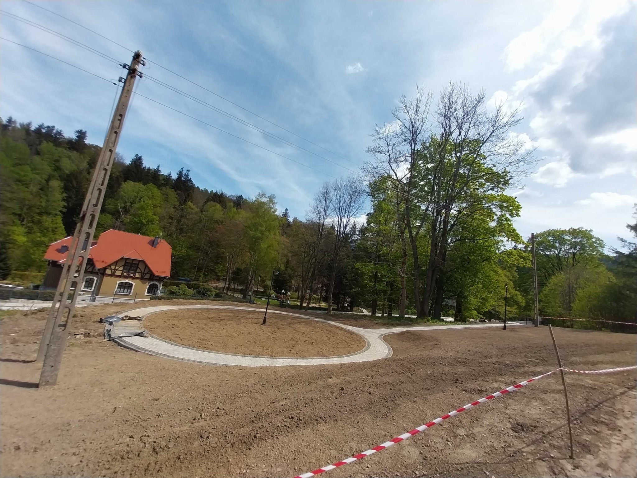 Kształtowanie terenu z wytyczonymi ścieżkami z kostki brukowej, przygotowane pod nasadzenia. W tle budynek z czerwoną dachówką i las.