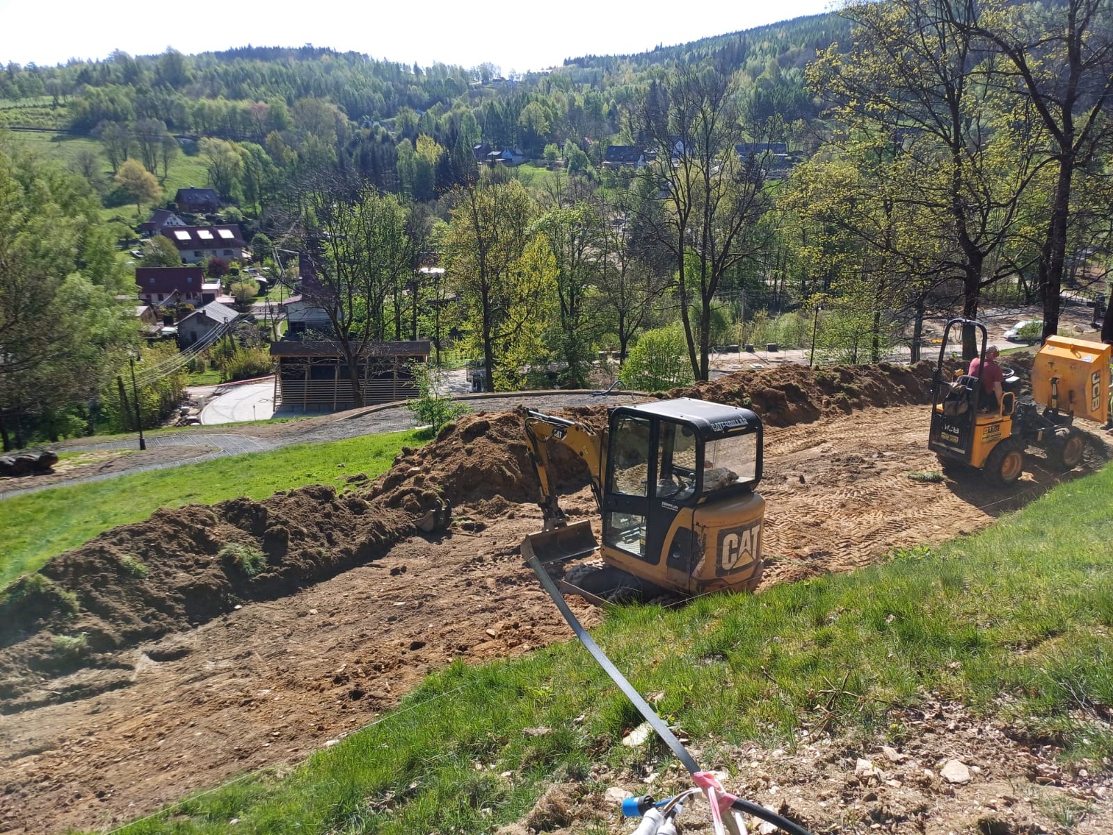 Żółty mini-koparka CAT i mniejsza koparka JCB na zboczu, w tle wieś i las. Wykopany rów w ziemi, przygotowania do prac ziemnych na nierównym terenie.