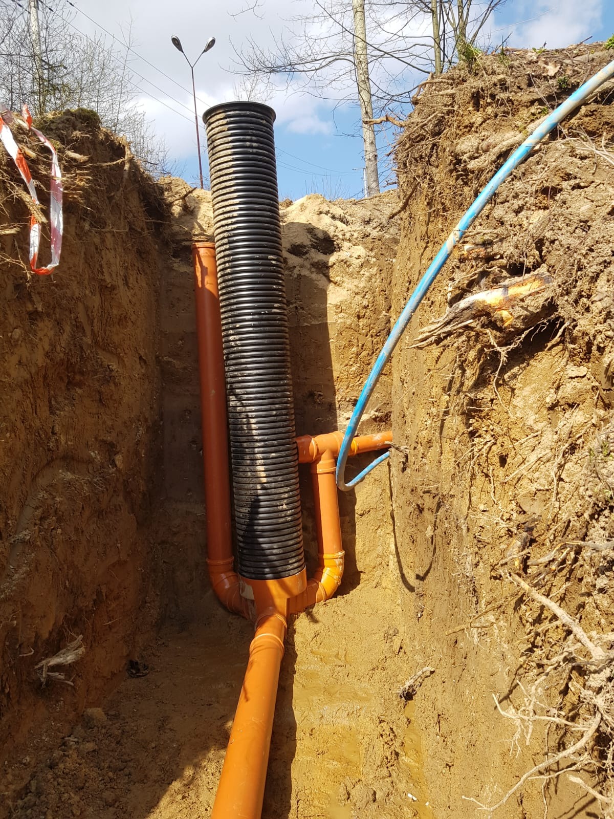 Głęboki wykop z ułożonym systemem rur drenarskich w Mirsku. Widoczne pomarańczowe rury połączone z czarną rurą karbowaną. Nad wykopem wystają korzenie drzew.