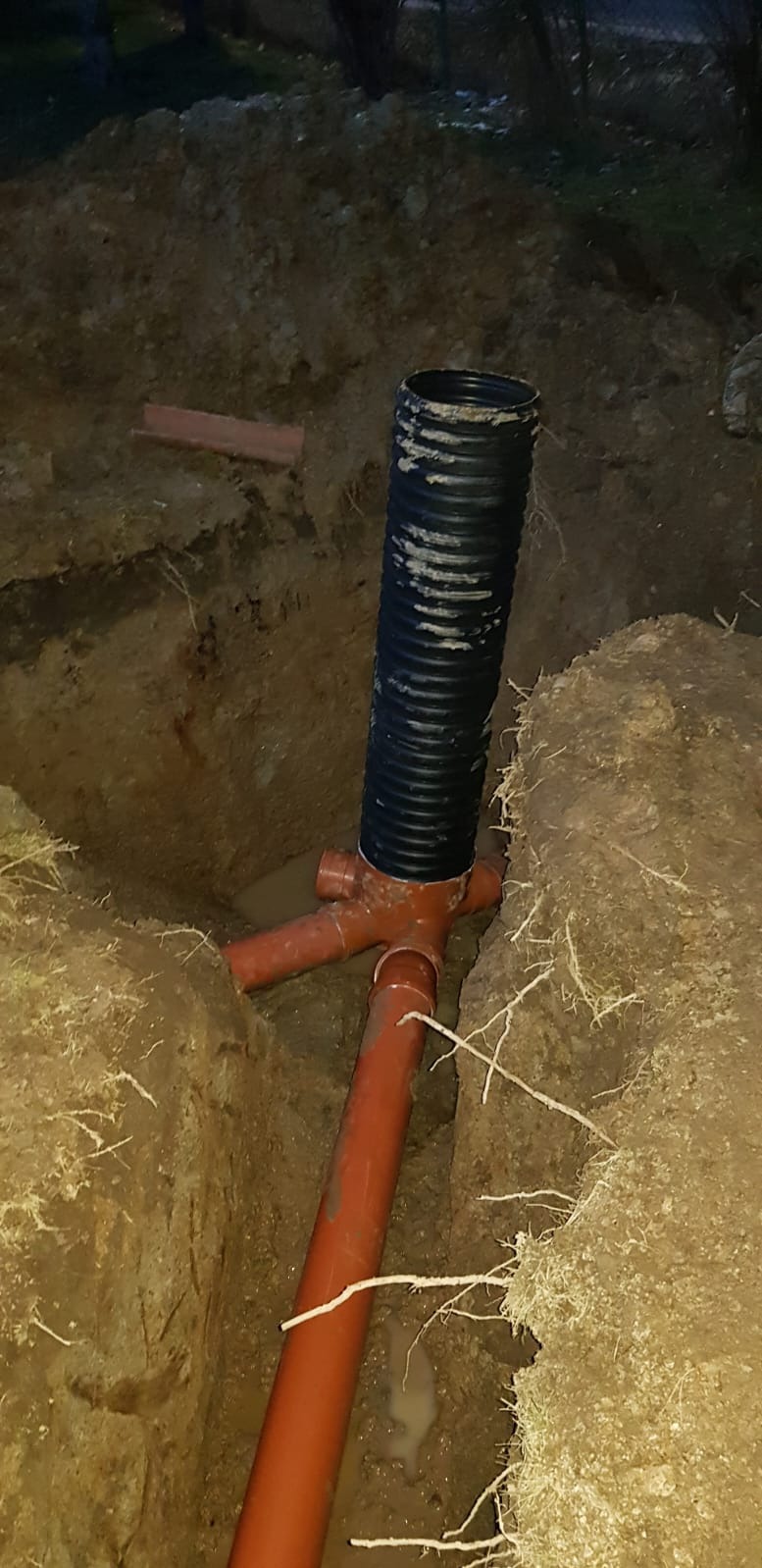 Instalacja drenażowa z rurą karbowaną w wykopie. Widoczne złącze trójnikowe i fragmenty rur. Ziemia wokół wykopu, słabe oświetlenie.