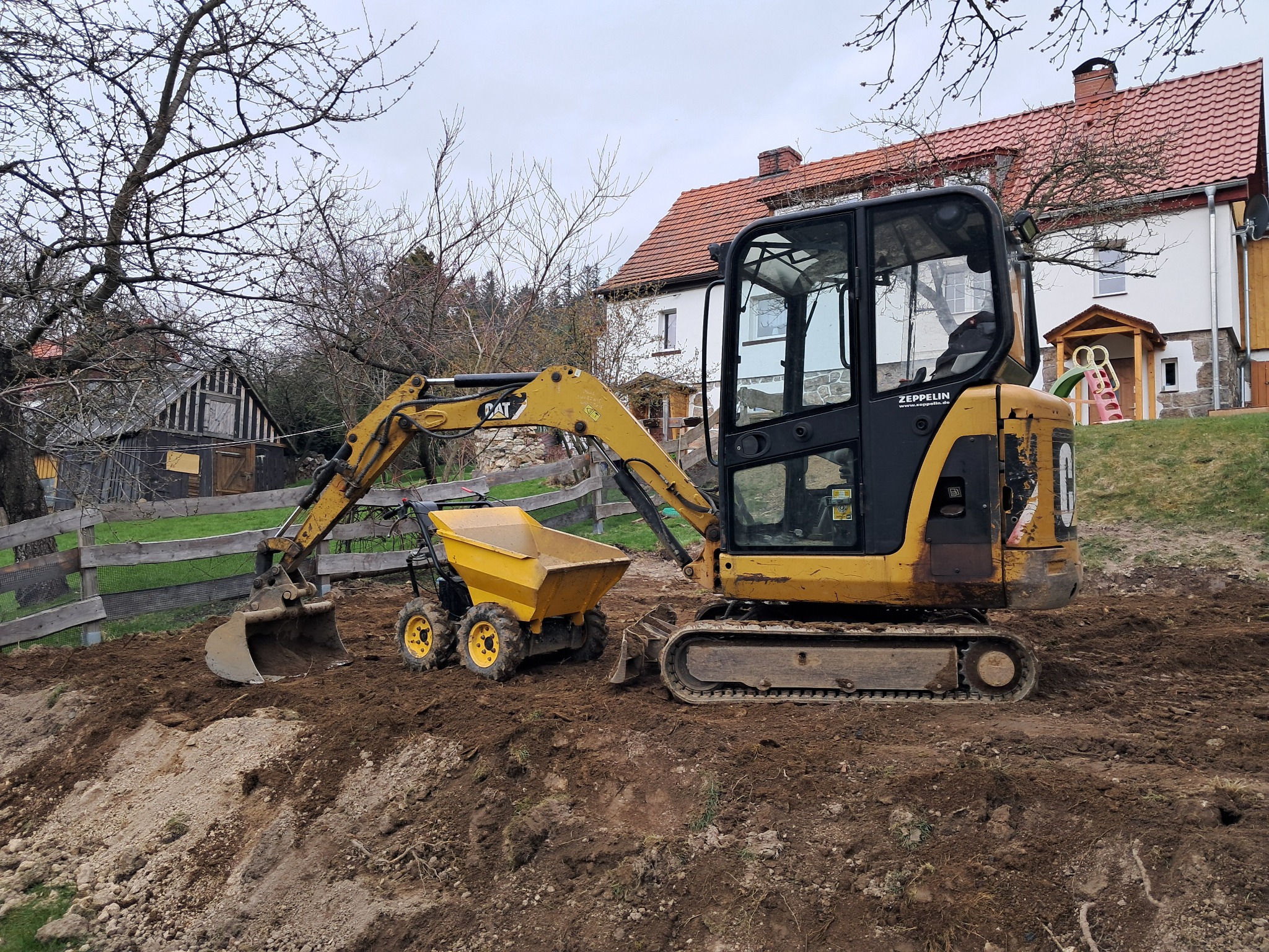Żółta mini koparka CAT i wywrotka na placu budowy, dom w tle. Widoczne ślady pracy w ziemi. Sprzęt Zeppelin.
