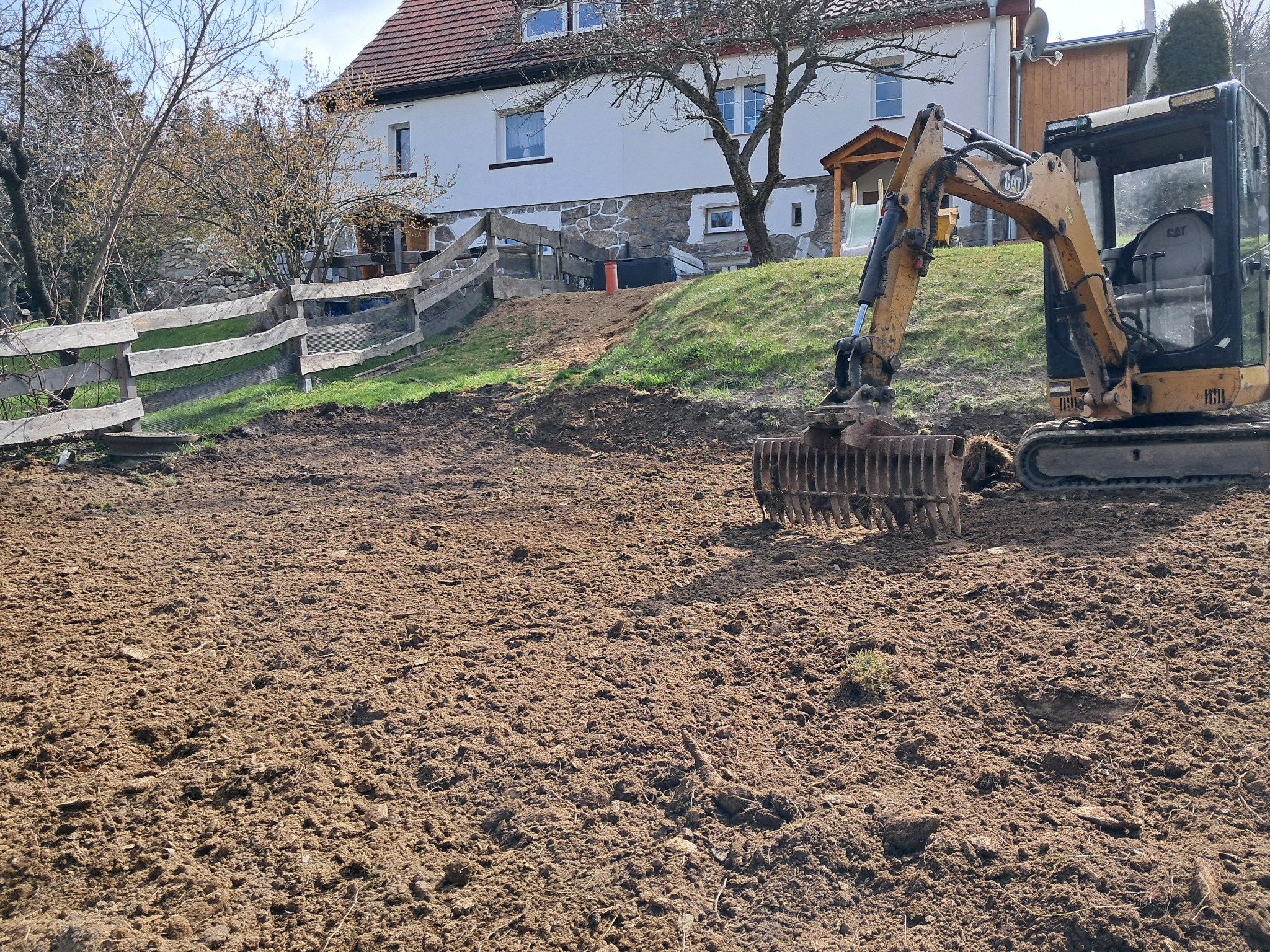 Żółta koparka CAT na świeżo przekopanym, brązowym terenie z widokiem na biały dom z kamienną podmurówką i drewnianym płotem w tle.