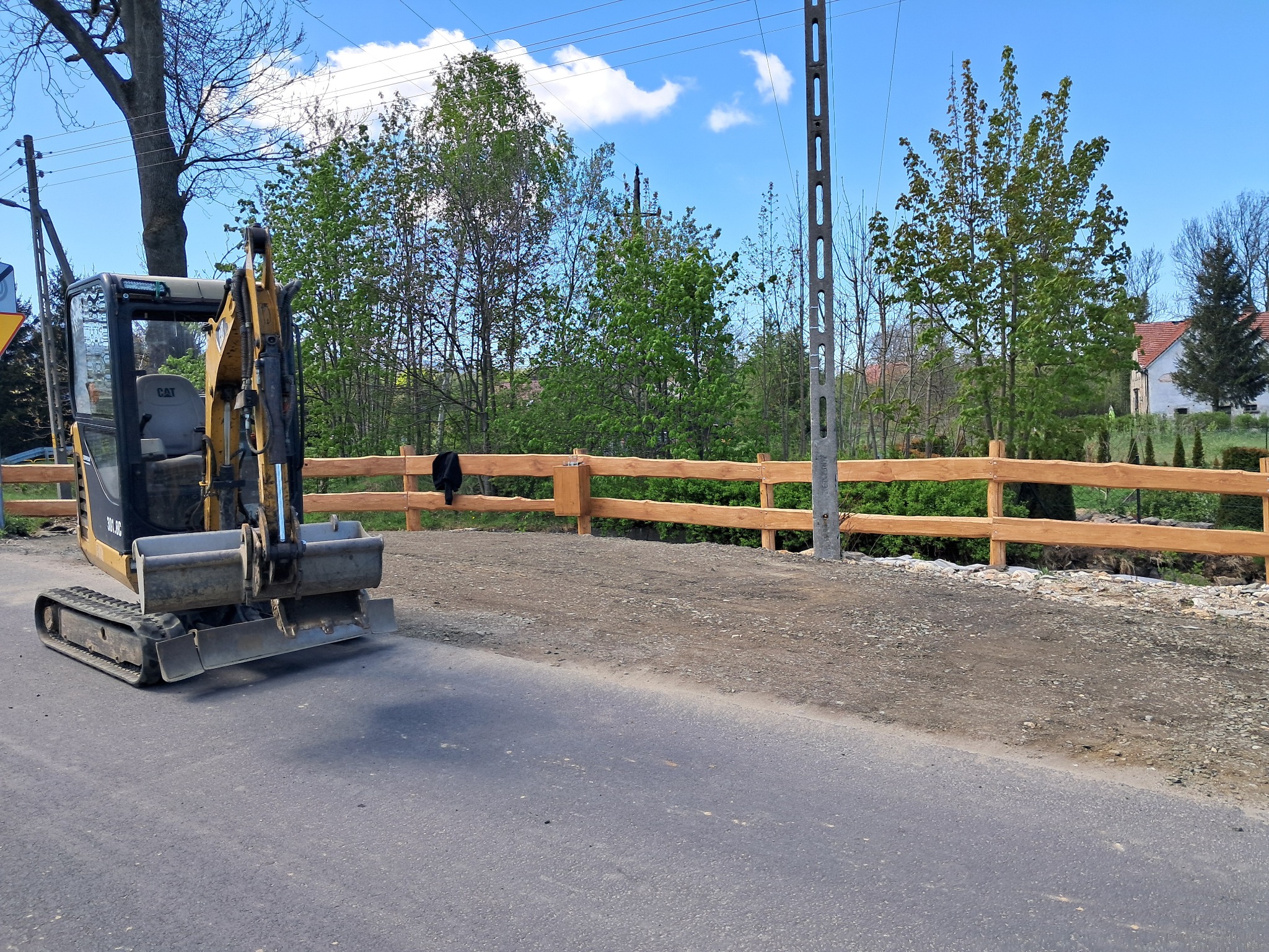 Żółta mini koparka CAT na tle drewnianego płotu, częściowo utwardzonego terenu i zieleni. Prace ziemne w toku, widok na krajobraz.