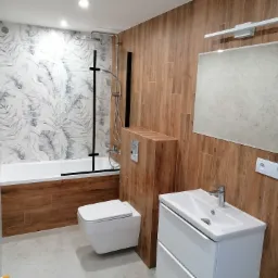 Nowoczesna łazienka po remoncie: drewnopodobne płytki, biała armatura, motyw liści na ścianie przy wannie. Minimalistyczny design i funkcjonalność.