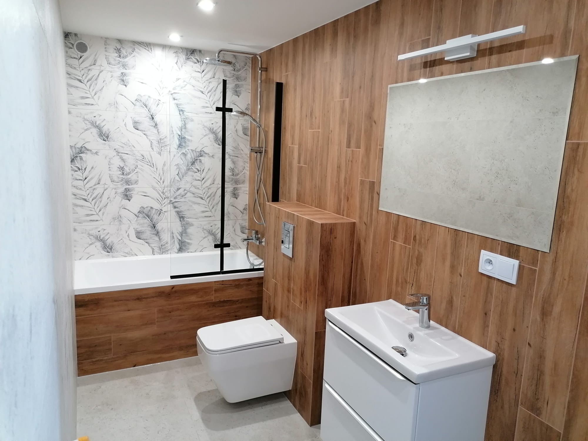 Nowoczesna łazienka po remoncie: drewnopodobne płytki, biała armatura, motyw liści na ścianie przy wannie. Minimalistyczny design i funkcjonalność.