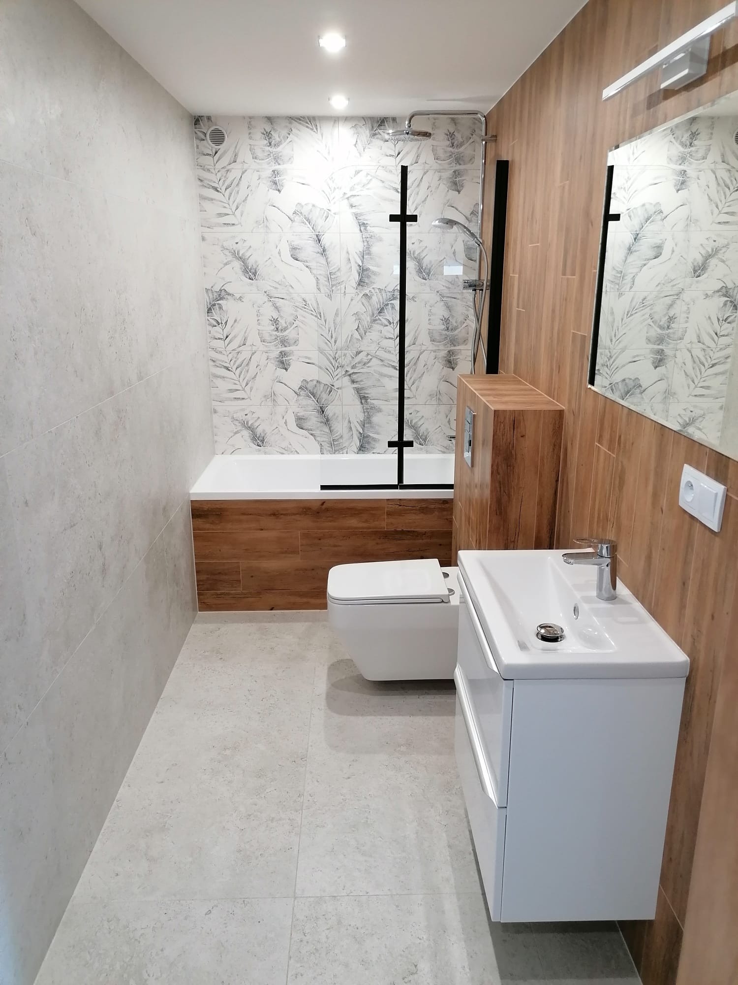 Nowoczesna łazienka po remoncie: biała umywalka z szafką, toaleta w zabudowie, wanna z parawanem i płytki z motywem liści na ścianie. Drewnopodobne akcenty.
