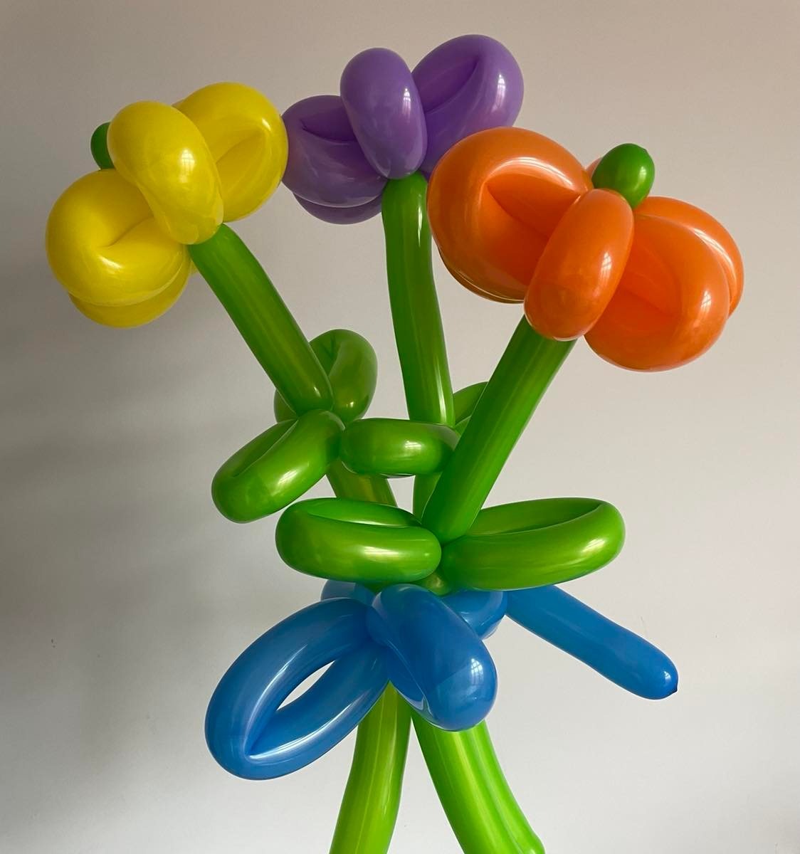 Bukiet kwiatów uformowany z kolorowych balonów: żółty, fioletowy, pomarańczowy, zielony i niebieski, na jednolitym, jasnym tle.