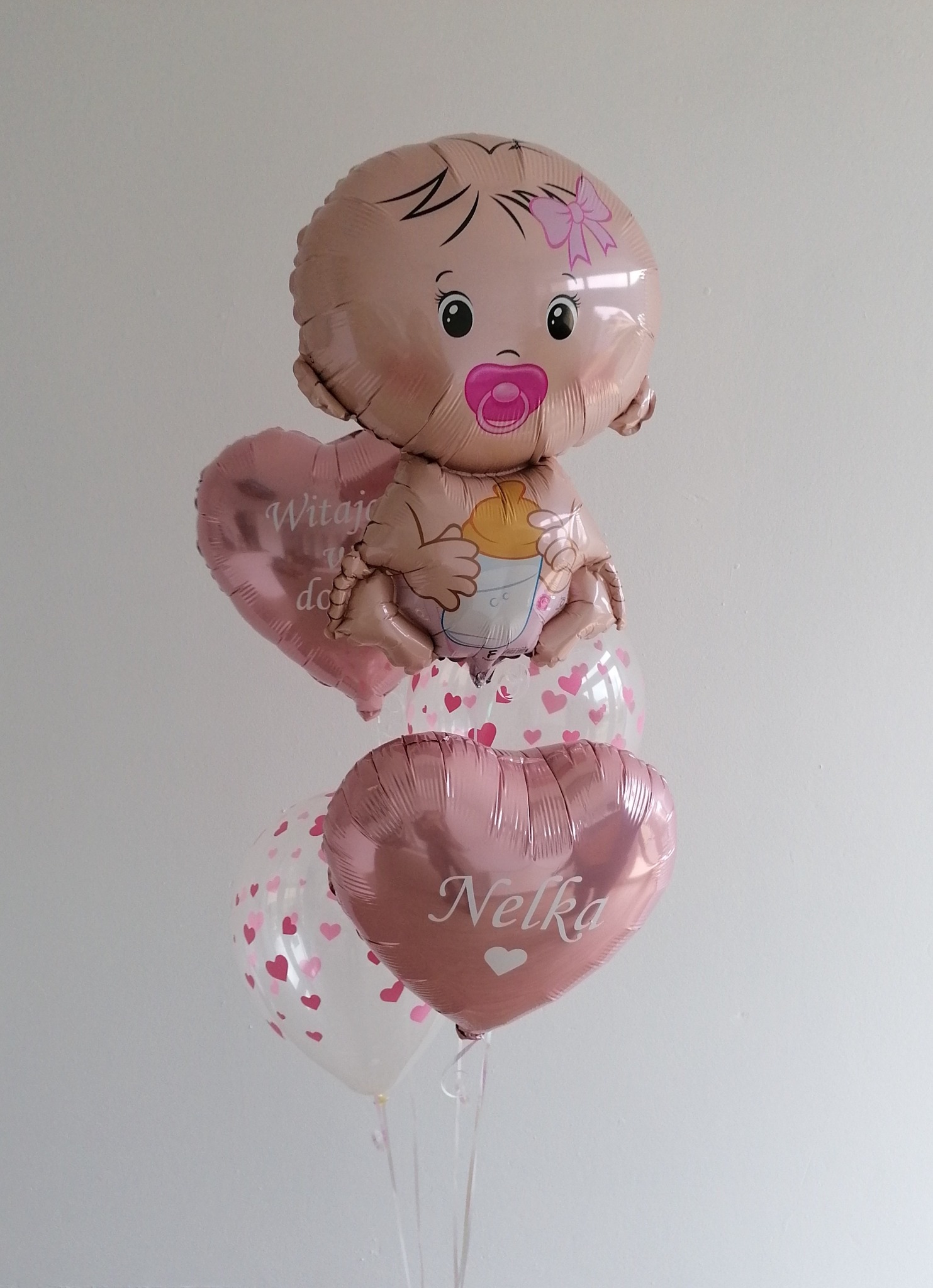 Balon foliowy w kształcie niemowlaka z butelką, unoszący się nad dwoma balonami w kształcie serc, z których jeden ma napis 'Witaj w domu', a drugi imię 'Nelka', na tle białej ściany.