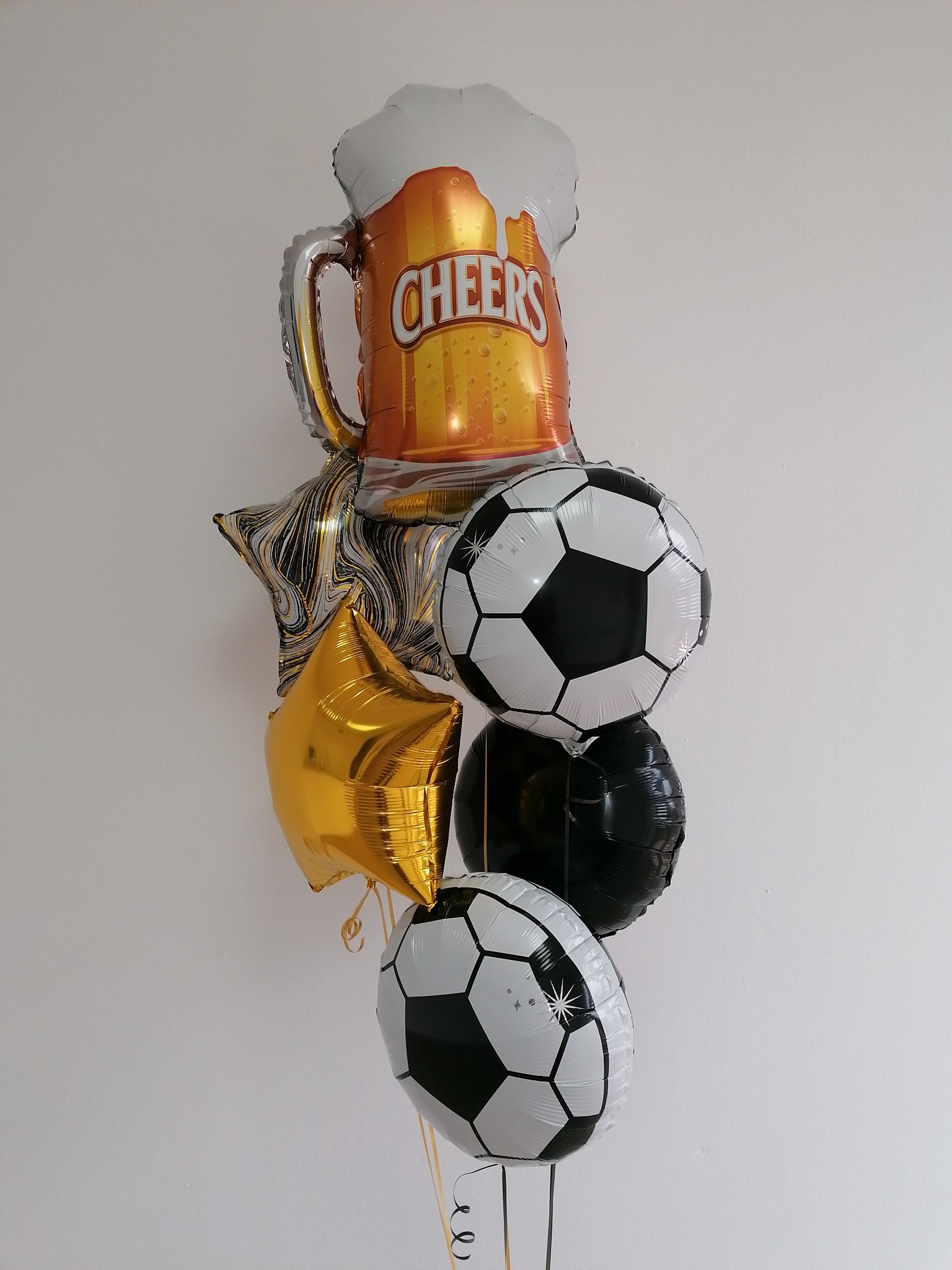Bukiet balonów foliowych: kufla piwa z napisem Cheers, dwóch piłek nożnych, złotej gwiazdy i czarnego balonu, zawieszonych na tle białej ściany.