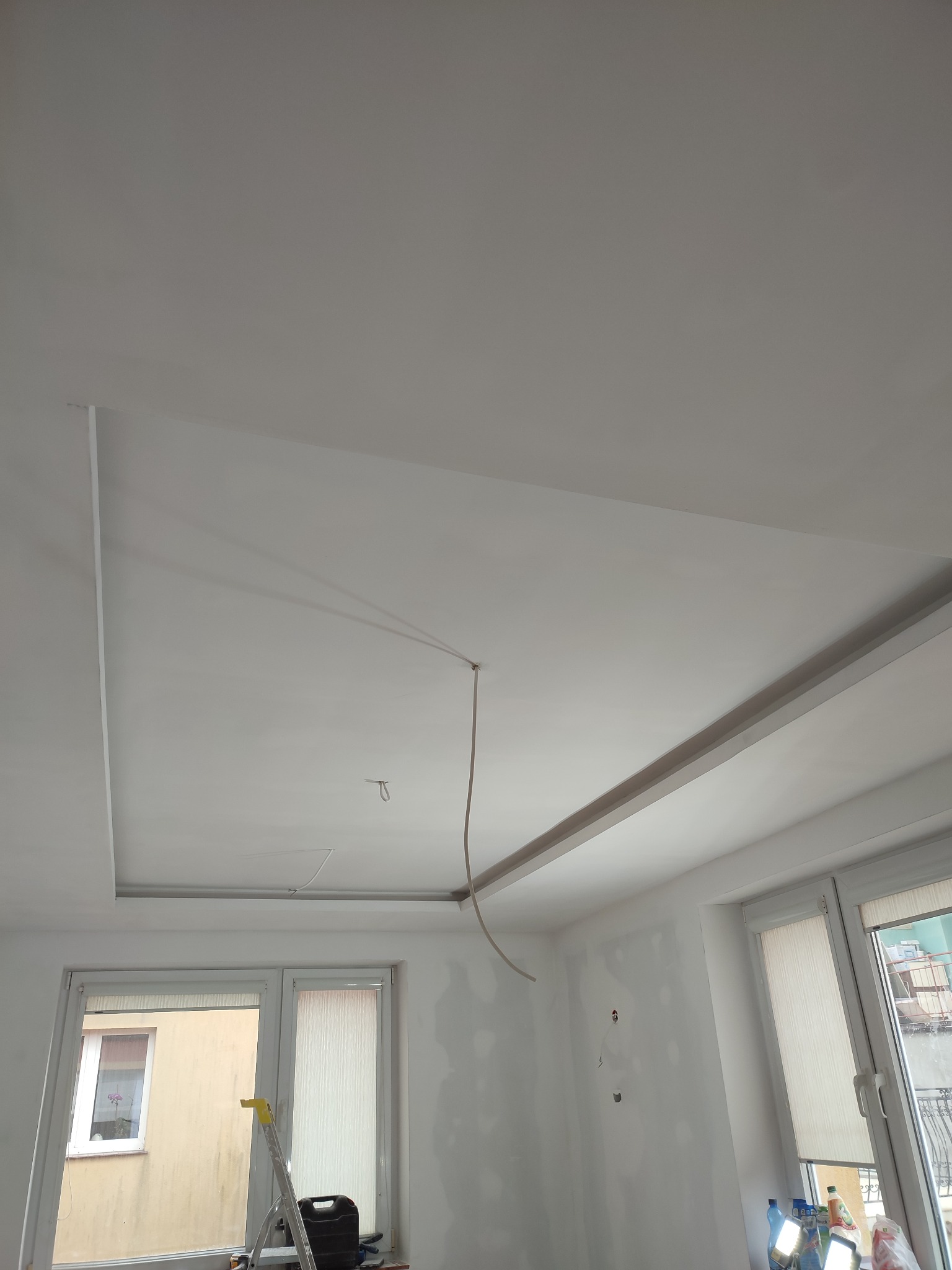 Wnętrze pokoju w trakcie wykańczania, widoczny fragment sufitu podwieszanego z elementami oświetlenia, ściany przygotowane do malowania z widocznymi śladami po szpachlowaniu, drabina oparta o okno.