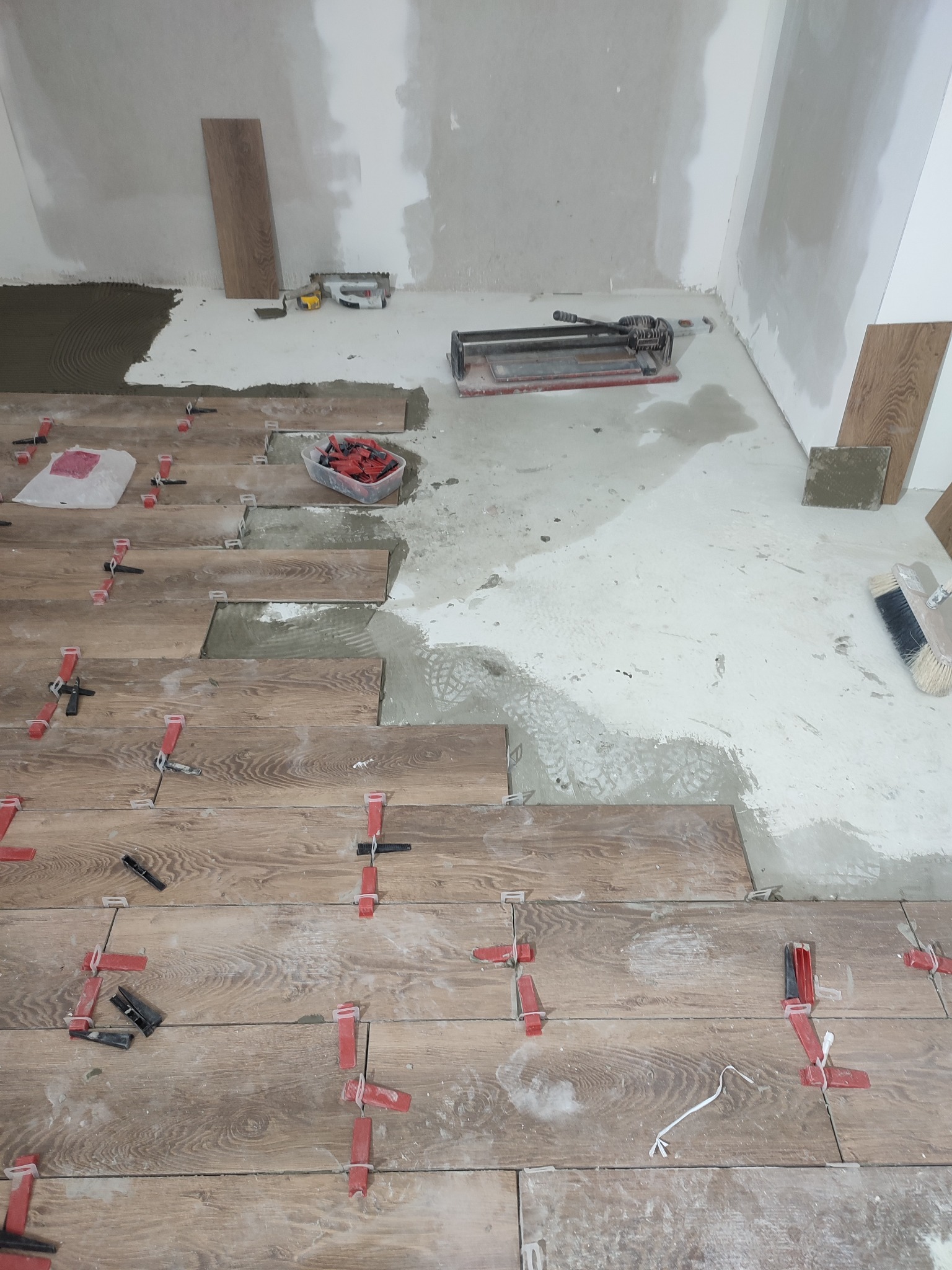Układanie podłogi z płytek imitujących drewno, z użyciem systemu poziomowania płytek (czerwone klipsy i czarne kliny), widoczna zaprawa klejowa, narzędzia glazurnicze i płyta do cięcia płytek