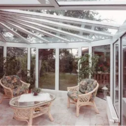 Jasna oranżeria z białymi ramami, szklanym dachem i ścianami, rattanowe meble wypoczynkowe, widok na ogród.