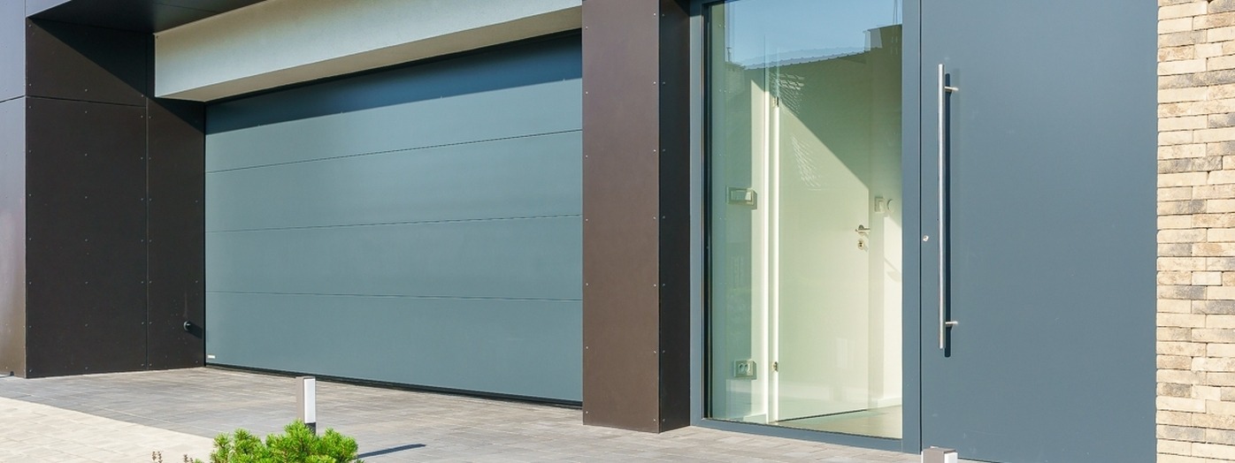 Nowoczesna elewacja domu z szarą segmentową bramą garażową, przeszklonymi drzwiami wejściowymi z długim uchwytem i dekoracyjną ścianą z kamienia.