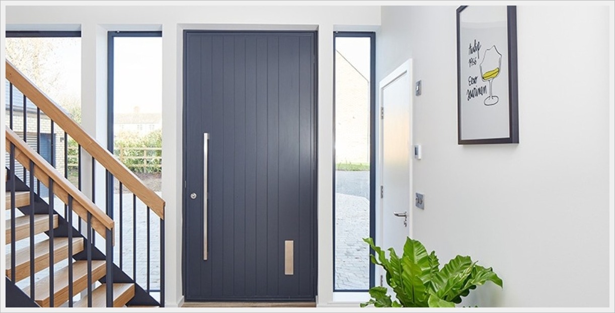Nowoczesne wejście do domu z ciemnoszarymi drzwiami zewnętrznymi z pionowymi panelami i długim, pionowym uchwytem, widoczne schody z drewnianymi poręczami i czarnymi balustradami, obok drzwi wąskie...