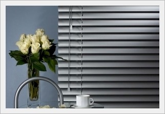 Szare poziome żaluzje okienne z metalicznym połyskiem, częściowo zasłonięte, w tle wazon z białymi różami i biała filiżanka na stoliku z chromowanym wykończeniem.