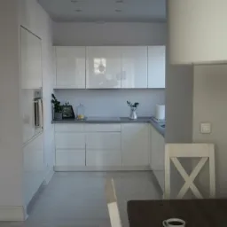 Nowoczesna, minimalistyczna kuchnia z białymi szafkami, szarym blatem i drewnianym stołem z filiżanką kawy; krzesło z oparciem w kształcie litery X.