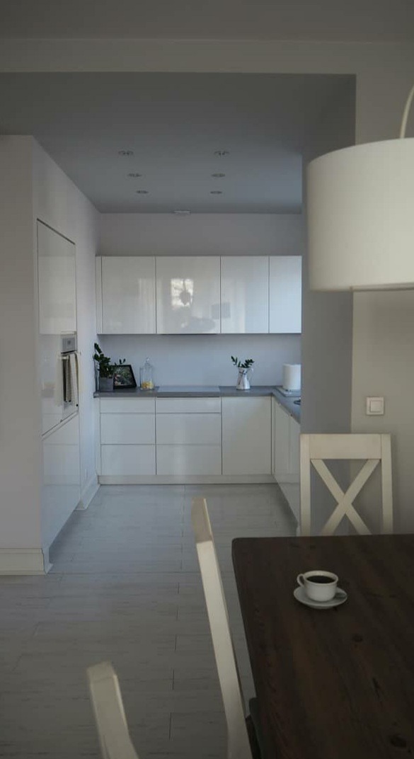 Nowoczesna, minimalistyczna kuchnia z białymi szafkami, szarym blatem i drewnianym stołem z filiżanką kawy; krzesło z oparciem w kształcie litery X.