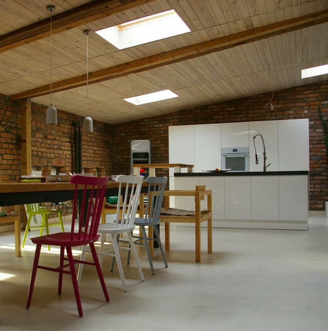 Wnętrze loftu z ceglaną ścianą, drewnianym sufitem z oknami dachowymi, białą zabudową kuchenną i stołem z kolorowymi krzesłami.