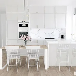 Jasna, minimalistyczna kuchnia z białymi szafkami, marmurowym backsplash'em, drewnianym stołem i białymi krzesłami w stylu skandynawskim, z bukietem różowych kwiatów na stole i trzema wiszącymi...
