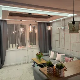 Nowoczesny salon z szarą sofą, drewnianym stolikiem kawowym i dekoracyjnymi lampami wiszącymi, z projektorem zamontowanym na ścianie.