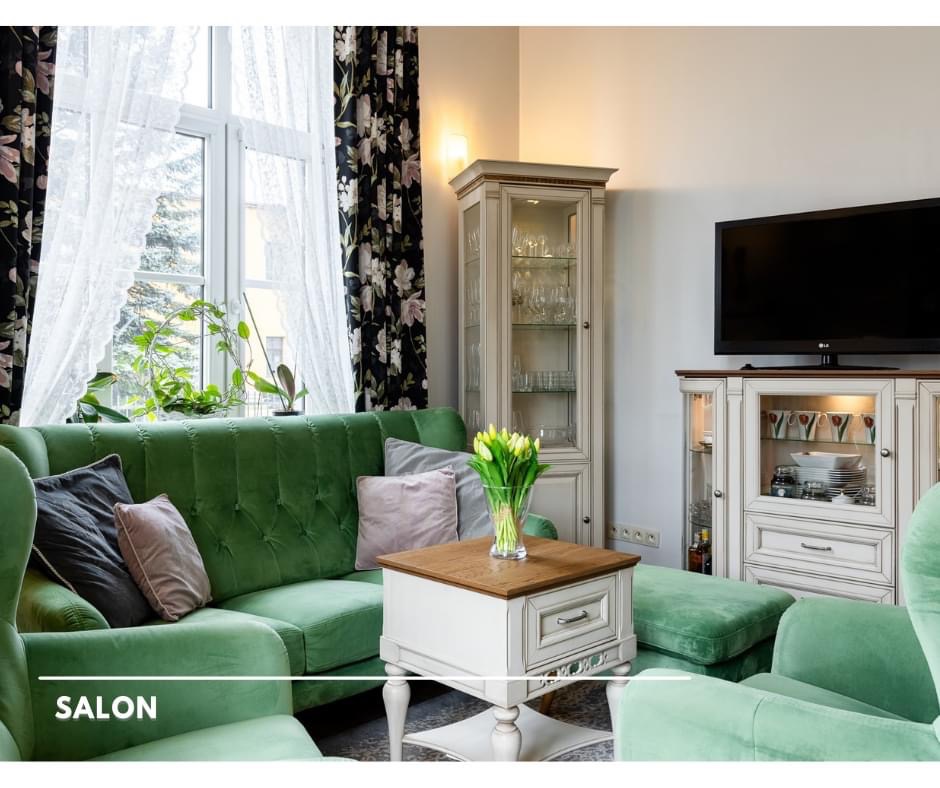 Jasnozielony salon z welurową sofą, stolikiem kawowym z bukietem tulipanów, witryną z zastawą i telewizorem, okno z kwiecistymi zasłonami.