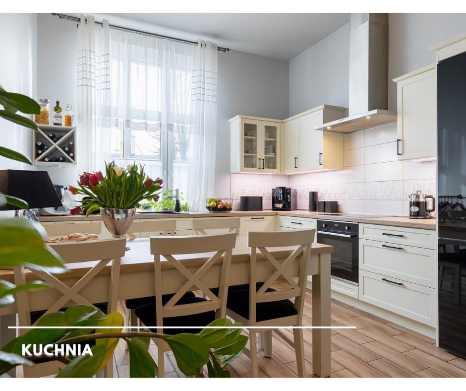 Jasna kuchnia w stylu klasycznym z drewnianym stołem i krzesłami, kwiatami w srebrnym wazonie i widokiem na okno z delikatnymi firankami.