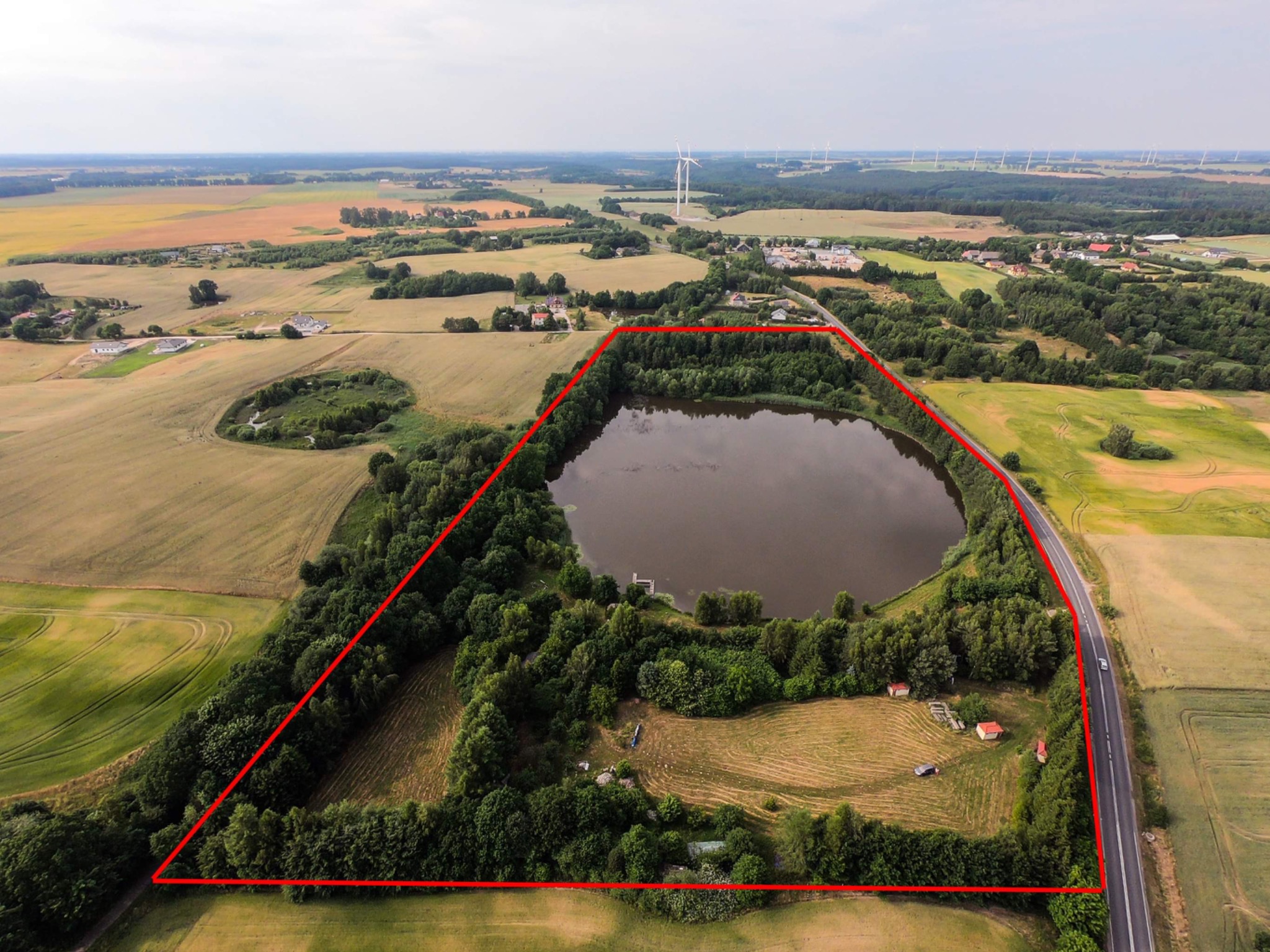 Widok z lotu ptaka na działkę z jeziorem otoczoną lasem i polami, wyznaczona czerwonym obrysem; w tle turbiny wiatrowe.