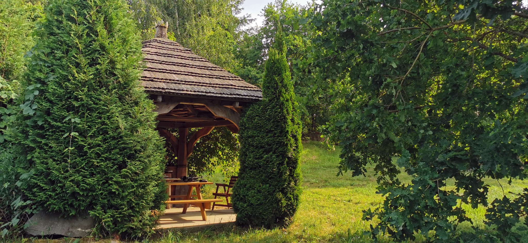 Drewniana altana z ławkami i stołem, otoczona zielenią drzew i krzewów, widok z zewnątrz.