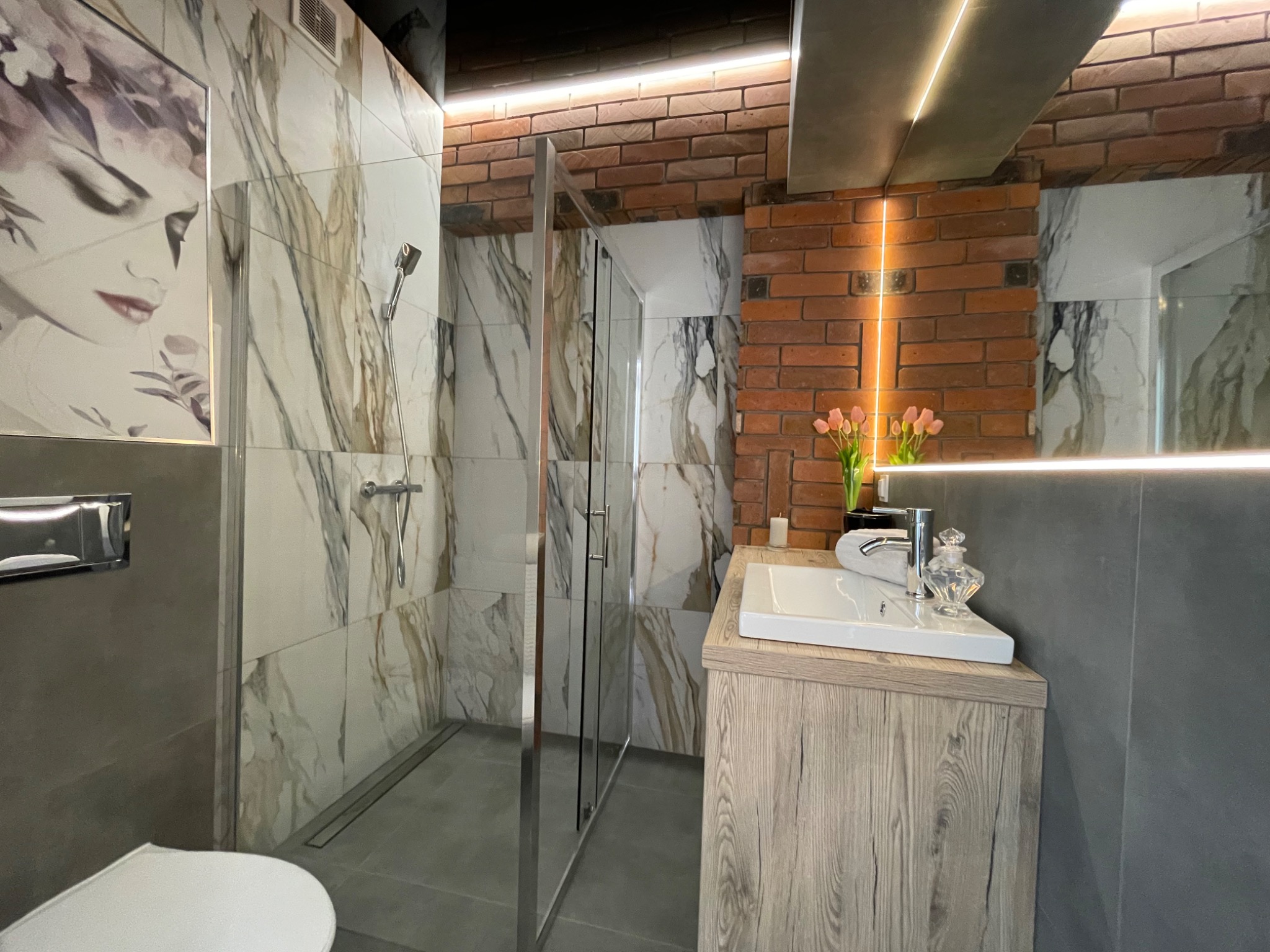 Nowoczesna łazienka z kabiną prysznicową typu walk-in, marmurowymi płytkami z motywem twarzy, ceglaną ścianą z oświetleniem LED, umywalką nablatową z drewnopodobną szafką i dekoracyjnym flakonem.
