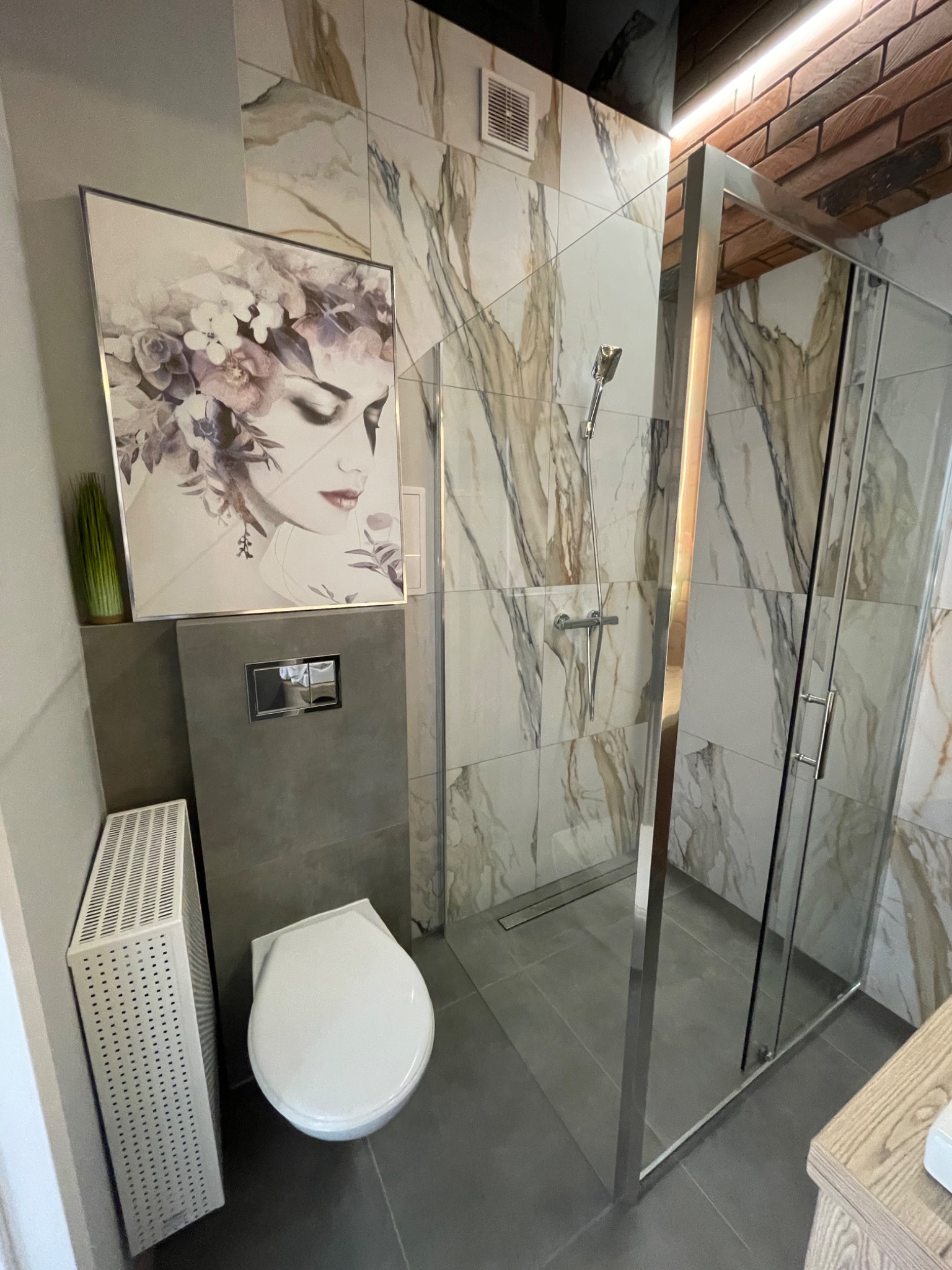Nowoczesna łazienka z kabiną prysznicową z chromowanymi profilami, marmurowe płytki ścienne, toaleta podwieszana, biały grzejnik żeberkowy i obraz z motywem kobiecym.