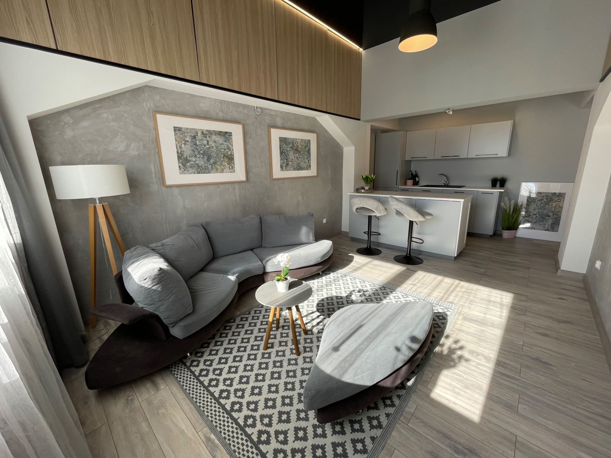 Jasne, przestronne wnętrze mieszkania z szarym narożnikiem, stolikiem kawowym z kwiatami, dywanem w geometryczne wzory i otwartą kuchnią z hokerami w tle.