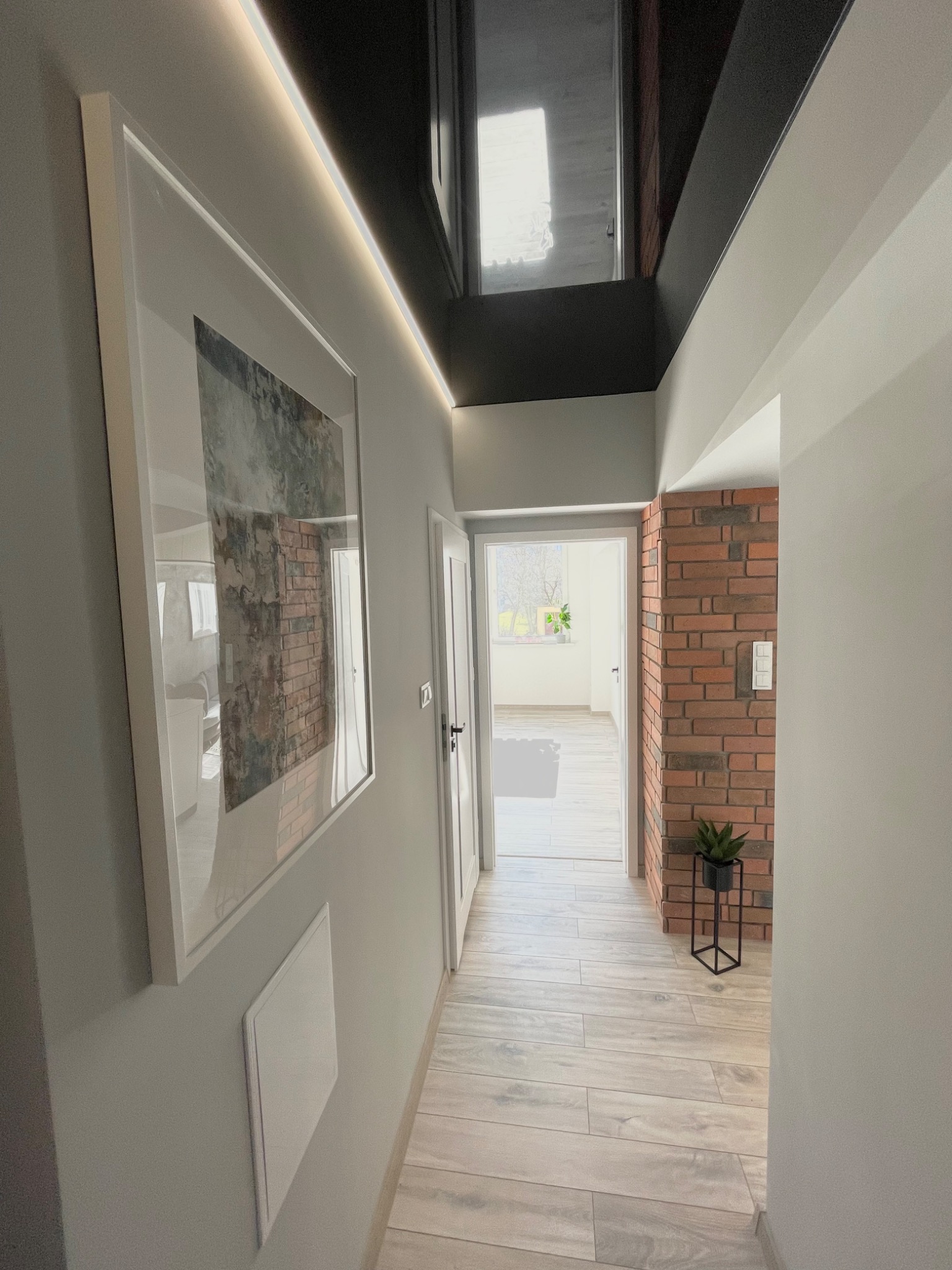 Wnętrze korytarza z drewnopodobną podłogą, ścianą z cegły, obrazem w białej ramie i podświetlanym sufitem w odcieniach szarości i bieli.