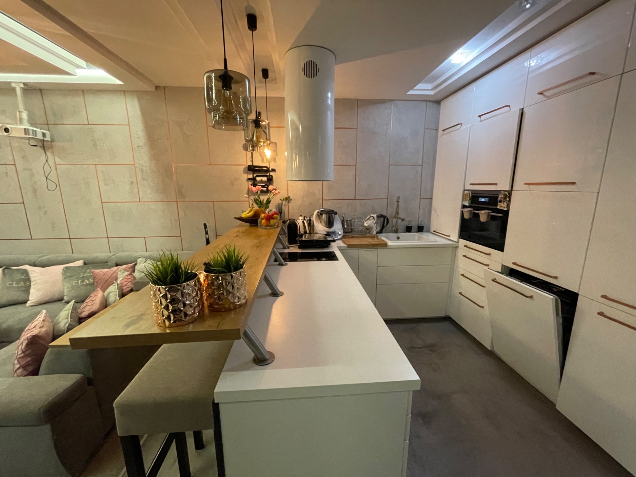 Nowoczesna kuchnia z białymi, lakierowanymi szafkami, drewnianym blatem i wyspą, połączona z salonem, z dekoracyjnymi lampami wiszącymi i roślinami w ozdobnych doniczkach.